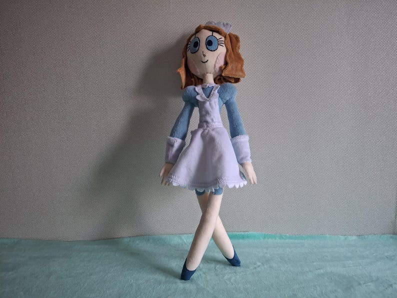 May include: A handmade doll in a maid outfit. The doll has brown hair, blue eyes, and a light skin tone. The outfit is light blue and white, with a white apron and a matching bonnet. The doll is standing with its legs crossed and wearing blue shoes.