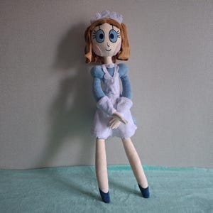 May include: A handmade doll dressed in a blue and white outfit, reminiscent of a maid's uniform. The doll has brown hair, blue eyes, and a white cap. The doll's legs are long and slender, with blue shoes. The doll is standing on a light blue surface.