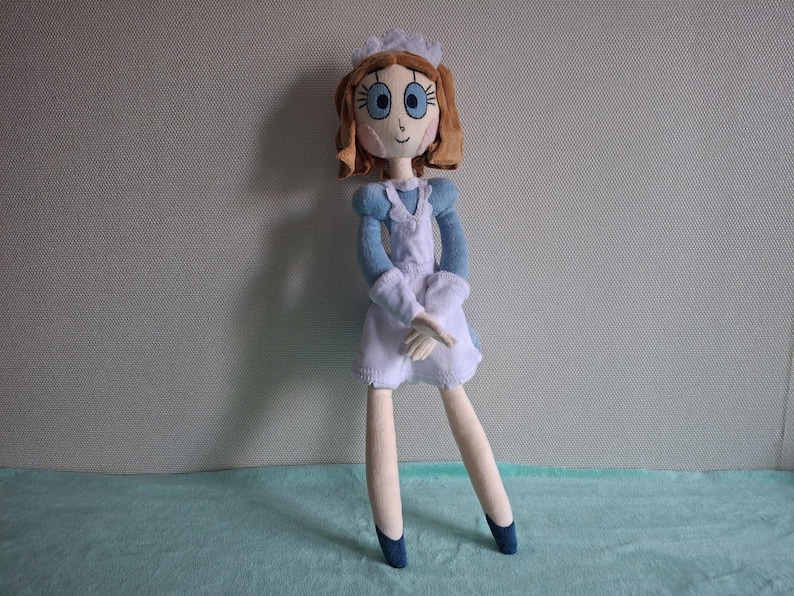 May include: A handmade doll dressed in a blue and white outfit, reminiscent of a maid's uniform. The doll has brown hair, blue eyes, and a white cap. The doll's legs are long and slender, with blue shoes. The doll is standing on a light blue surface.