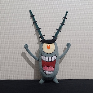 Op de afbeelding: Een groen pluche speelgoed van Plankton uit de cartoon Spongebob Squarepants. Plankton is een klein, eenogig wezen met een grote mond en een paar antennes.