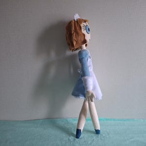 May include: A handmade doll with a brown head, blue eyes, and a blue and white dress. The doll has a white cap and is standing on a light blue surface. The doll's legs are made of a light-colored material, and it is wearing blue shoes.