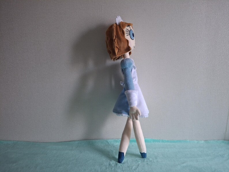 May include: A handmade doll with a brown head, blue eyes, and a blue and white dress. The doll has a white cap and is standing on a light blue surface. The doll's legs are made of a light-colored material, and it is wearing blue shoes.