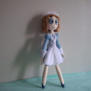 May include: A handmade doll dressed in a blue and white maid outfit. The doll has brown hair, blue eyes, and a white cap. The doll's arms and legs are thin and the doll is standing on a light blue surface.