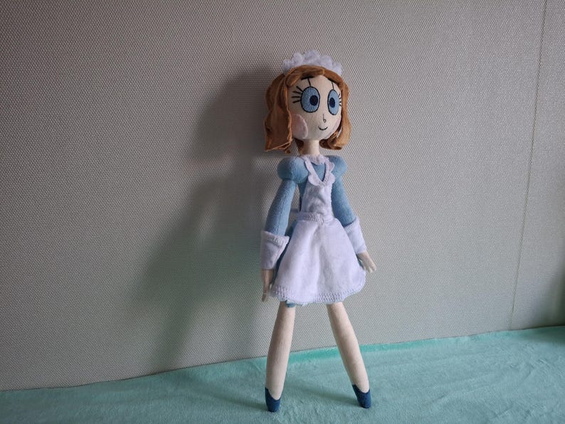 May include: A handmade doll dressed in a blue and white maid outfit. The doll has brown hair, blue eyes, and a white cap. The doll's arms and legs are thin and the doll is standing on a light blue surface.