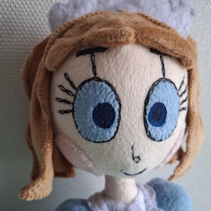 May include: A plush doll with a light skin tone, brown hair, and large blue eyes. The doll has a small nose and a stitched smile. It wears a blue and white outfit and a white cap. The doll is a handmade item.