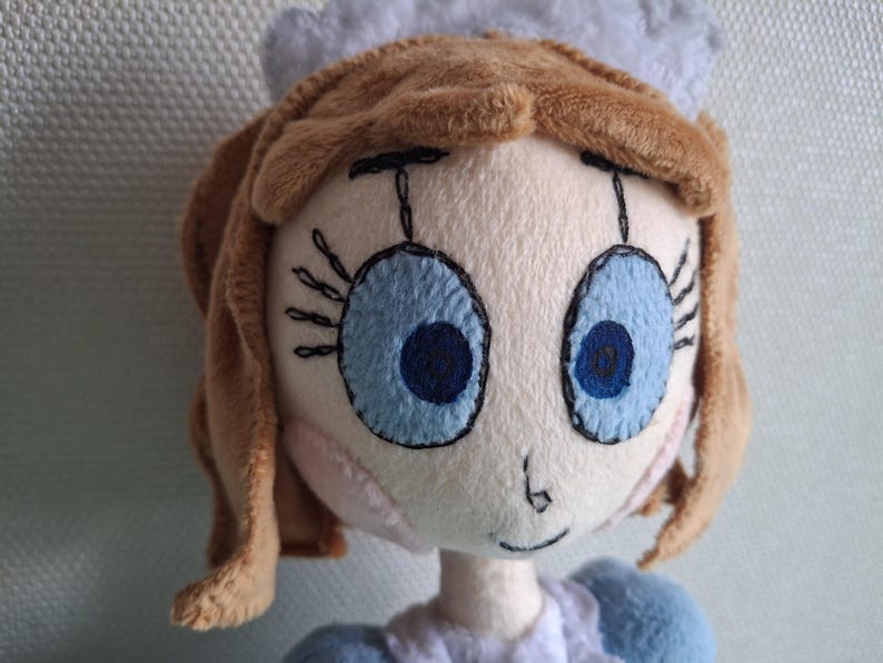 May include: A plush doll with a light skin tone, brown hair, and large blue eyes. The doll has a small nose and a stitched smile. It wears a blue and white outfit and a white cap. The doll is a handmade item.