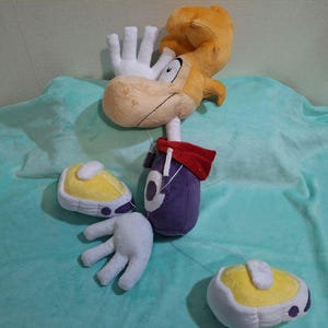 May include: A plush toy of the video game character Rayman, featuring a tan face and orange hair. The plush has white hands and feet, a purple body, and yellow shoes. A red cape is attached to the body.