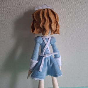 May include: A handmade doll, viewed from the back, dressed in a blue dress with white accents. The doll has brown, wavy hair and a white cap. The dress features a white apron-like design and a white belt. The doll's legs are a light beige color.