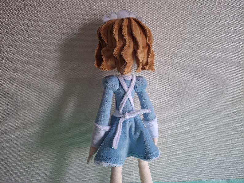 May include: A handmade doll, viewed from the back, dressed in a blue dress with white accents. The doll has brown, wavy hair and a white cap. The dress features a white apron-like design and a white belt. The doll's legs are a light beige color.