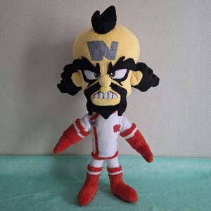 Puede incluir: Un muñeco de peluche de un personaje con una cabeza grande de color amarillo, cabello negro y una barba negra prominente. El personaje tiene una letra N gris en la frente, viste una camisa blanca de manga larga con detalles rojos, guantes rojos y botas rojas.