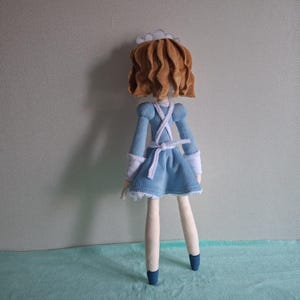 May include: A handmade doll wearing a light blue dress with white trim and a white apron. The doll has brown hair and is wearing a white cap. The doll's legs are cream-colored, and it is wearing blue shoes.