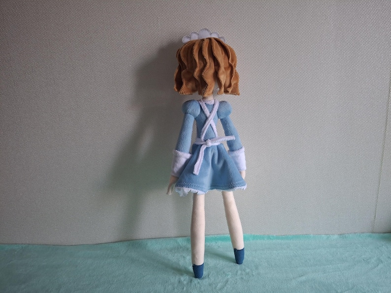 May include: A handmade doll wearing a light blue dress with white trim and a white apron. The doll has brown hair and is wearing a white cap. The doll's legs are cream-colored, and it is wearing blue shoes.