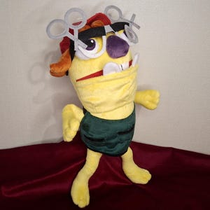 Puede incluir: Un juguete de peluche amarillo con un chaleco verde y un par de gafas plateadas. El juguete tiene una cabeza roja y amarilla con una nariz morada y una amplia sonrisa.