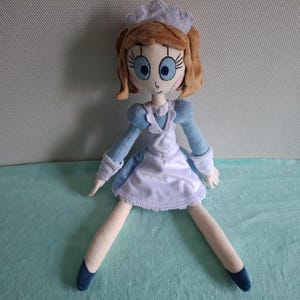 May include: A handmade doll with a light blue and white dress, a white apron, and a matching cap. The doll has brown hair, large blue eyes, and blue shoes. The doll is sitting with its legs spread.