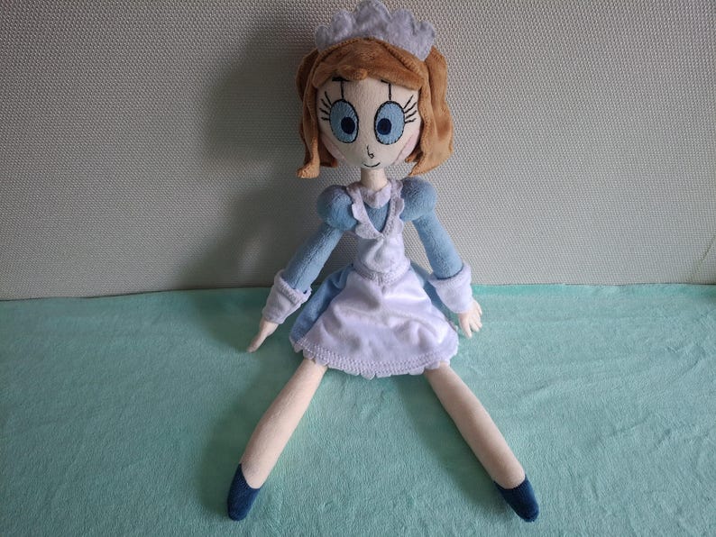 May include: A handmade doll with a light blue and white dress, a white apron, and a matching cap. The doll has brown hair, large blue eyes, and blue shoes. The doll is sitting with its legs spread.
