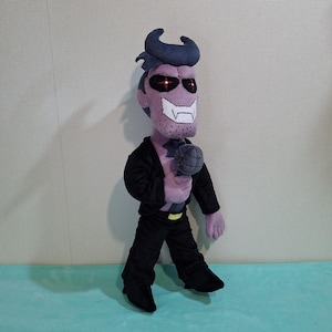 May include: A plush toy of a purple-skinned character with black hair, wearing a black suit jacket and black pants. The character has red eyes and a mischievous grin. The toy is holding a black microphone.
