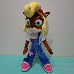 May include: A plush doll of a cartoon character with brown fur, green eyes, and yellow hair. The doll wears a white shirt, blue overalls, and pink shoes. A pink bow is on the doll's head.