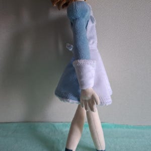 May include: A plush doll wearing a light blue and white dress with long sleeves. The doll has light skin, blonde hair, and is standing on a light blue surface. The doll's dress has white cuffs and a white collar.