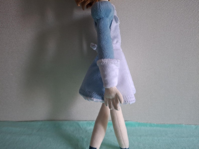 May include: A plush doll wearing a light blue and white dress with long sleeves. The doll has light skin, blonde hair, and is standing on a light blue surface. The doll's dress has white cuffs and a white collar.