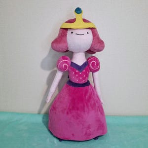Peut inclure: Une poupée en peluche rose de la princesse Bubblegum du dessin animé Adventure Time. Elle porte une robe rose avec un corsage violet et une couronne jaune avec un bijou bleu sur le dessus.