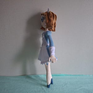 May include: A handmade doll featuring a character with light skin, blue eyes, and auburn hair. The doll wears a blue dress with white accents, a white apron, and blue shoes. The doll is standing on a light blue surface.