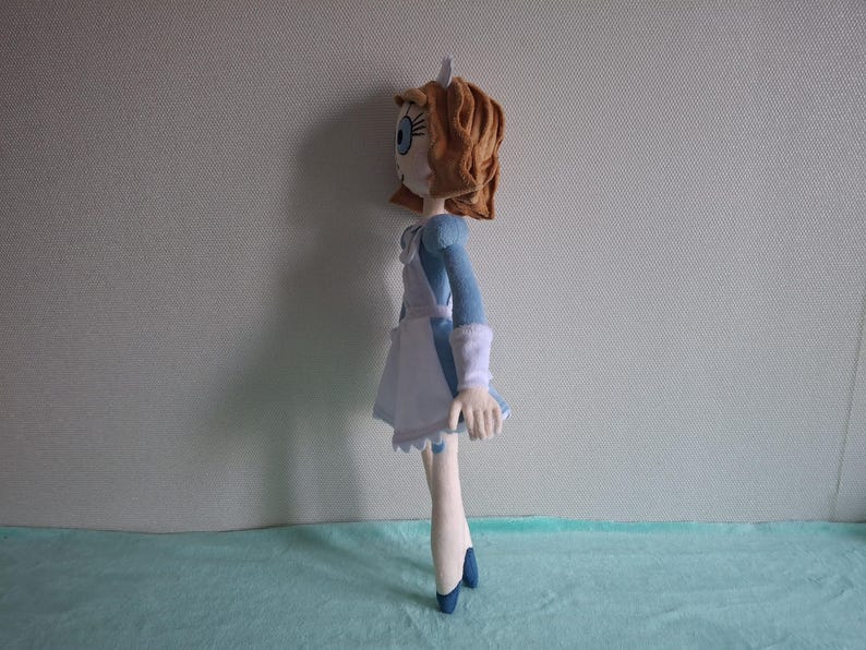 May include: A handmade doll featuring a character with light skin, blue eyes, and auburn hair. The doll wears a blue dress with white accents, a white apron, and blue shoes. The doll is standing on a light blue surface.