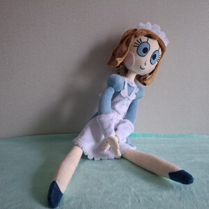 May include: A handmade doll with brown hair, blue eyes, and rosy cheeks. The doll is dressed in a blue and white outfit with a frilly white apron and a matching cap. The doll's legs are extended, and it has blue shoes.
