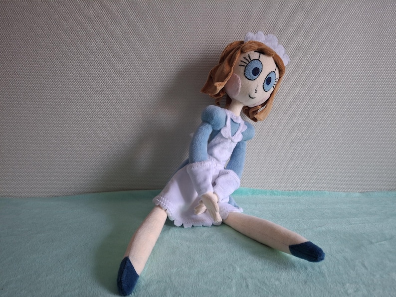 May include: A handmade doll with brown hair, blue eyes, and rosy cheeks. The doll is dressed in a blue and white outfit with a frilly white apron and a matching cap. The doll's legs are extended, and it has blue shoes.