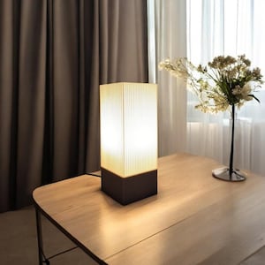 Puede incluir: Una lámpara de mesa rectangular moderna con una pantalla de color marrón claro con nervaduras y una base de color marrón oscuro. La lámpara está encendida e ilumina la superficie de la mesa.