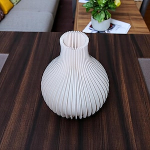 Può includere: Un vaso decorativo bianco con un design a coste unico. Il vaso ha un corpo bulboso e un collo stretto, con linee verticali che creano una superficie strutturata. Il vaso è realizzato in un materiale di colore chiaro e poggia su una superficie di legno scuro.