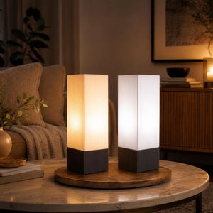 Pode incluir: Duas luminárias de mesa retangulares com cúpulas brancas caneladas e bases cinza-escuras. Uma luminária emite uma luz amarela quente, enquanto a outra emite uma luz branca fria. Ambas estão posicionadas lado a lado em uma bandeja de madeira.