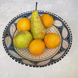 Puede incluir: Un cuenco de cerámica decorativo lleno de fruta fresca. El cuenco tiene un diseño de hojas azules y blancas alrededor del borde. La fruta incluye una manzana verde, una pera y varias naranjas. El cuenco está sobre un mantel blanco.