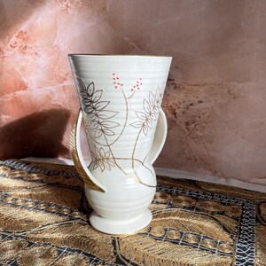Peut inclure: Un vase en céramique blanche avec des motifs floraux dorés et rouges et deux anses dorées. Le vase est posé sur une surface à motifs noirs et dorés.