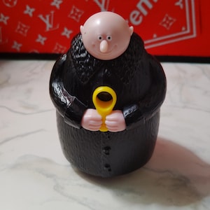 May include: A plastic figurine of a bald character in a black coat, holding a yellow object. The figure has a pale complexion and a distinctive nose. The background is a red surface with white lettering.
