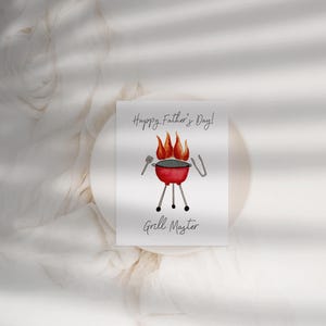 May include: A greeting card with the text "Happy Father's Day!" and "Grill Master." The card features a watercolour illustration of a red barbecue with flames, tongs, and a spatula. The card is on a white surface.