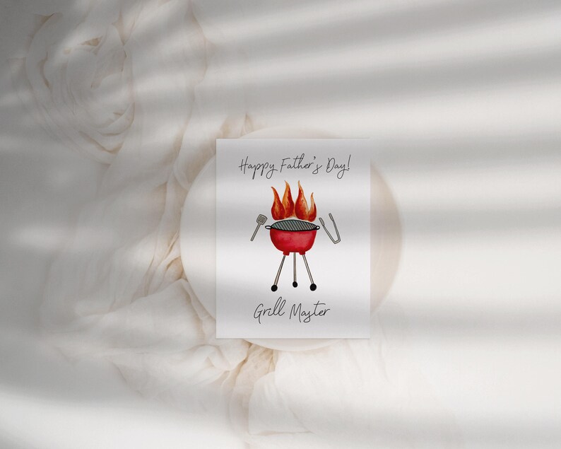 May include: A greeting card with the text "Happy Father's Day!" and "Grill Master." The card features a watercolour illustration of a red barbecue with flames, tongs, and a spatula. The card is on a white surface.