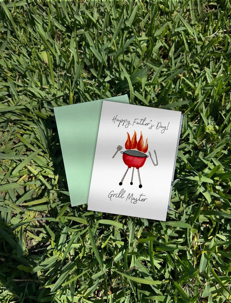 May include: A greeting card with a red barbecue illustration and the text "Happy Father's Day! Grill Master" is displayed on green grass. A pale green envelope is behind the card.
