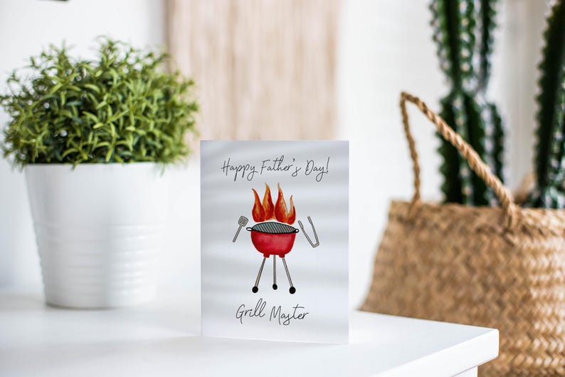 May include: A Father's Day card featuring a red barbecue illustration with flames, the text "Happy Father's Day!" and "Grill Master". The card is on a white surface, with a plant and a woven basket in the background.