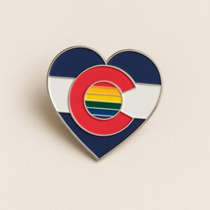 Può includere: Spilla a forma di cuore in smalto con il design della bandiera dello stato del Colorado. La spilla ha un bordo argentato e uno sfondo blu navy con una striscia bianca. Il centro presenta una "C" rossa con un cerchio arcobaleno all'interno.