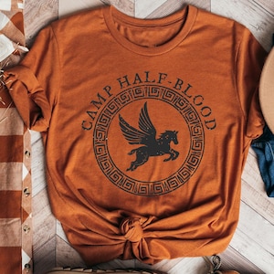 May include: A burnt orange t-shirt with the text "Camp Half-Blood" in a circle with a winged horse inside. The shirt is laid out on a wood floor with a brown and white plaid shirt, a pair of blue jeans, and a brown fedora hat.