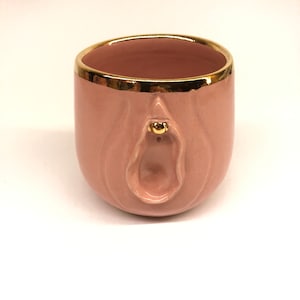 May include: A pink ceramic mug with a gold rim and a stylized vulva design on the front. The mug is a solid color with a smooth finish. The interior of the mug is also pink.