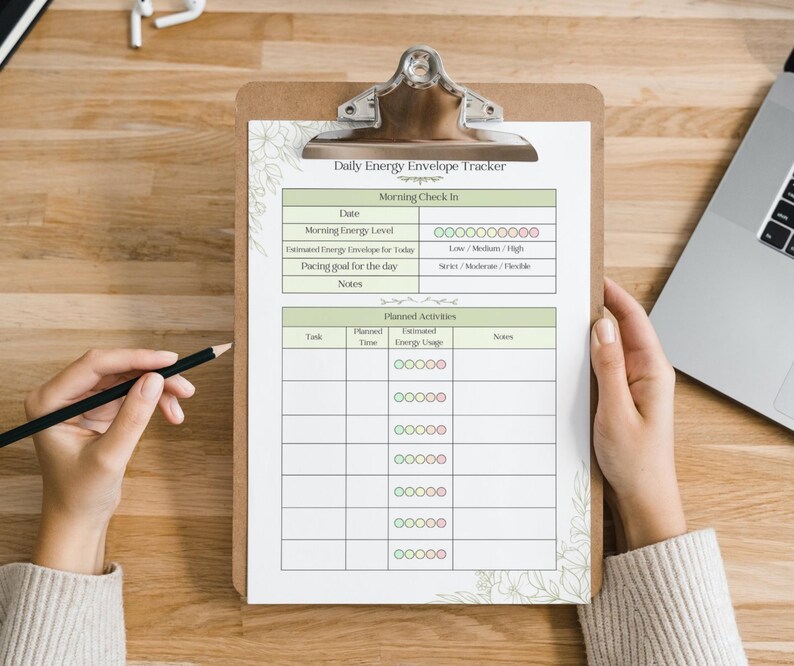 May include: A clipboard with a printed "Daily Energy Envelope Tracker" sheet. The sheet has sections for daily check-in and planned activities. A hand holds a pencil, ready to write. The clipboard is on a wooden surface.