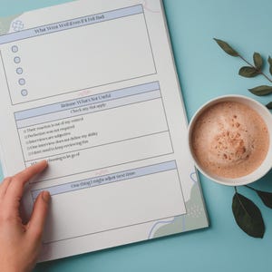 Puede incluir: Un diario con indicaciones para la autorreflexi&oacute;n, con secciones tituladas "Qu&eacute; sali&oacute; bien" y "Liberar lo que no es &uacute;til". Una taza de cer&aacute;mica blanca llena de una bebida espumosa se encuentra junto al diario.