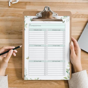 May include: A wooden clipboard with a printed goal-setting template. The template includes sections for 3-month, 6-month, 1-year, 5-year, 10-year, and 20-year goals. The design features a light green and white floral border. A hand holds a pencil.
