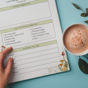 May include: A planner page with sections for tasks, challenges, and solutions, with the text "Low Energy." A hand points to a section labeled "Break It Into Tiny Steps." A mug of coffee sits nearby on a blue surface.
