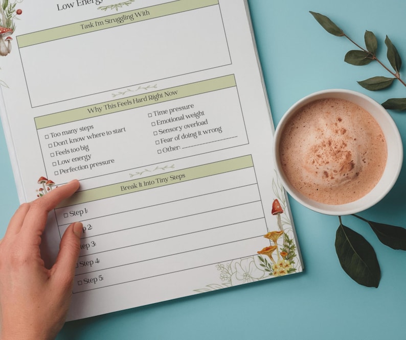 May include: A planner page with sections for tasks, challenges, and solutions, with the text "Low Energy." A hand points to a section labeled "Break It Into Tiny Steps." A mug of coffee sits nearby on a blue surface.