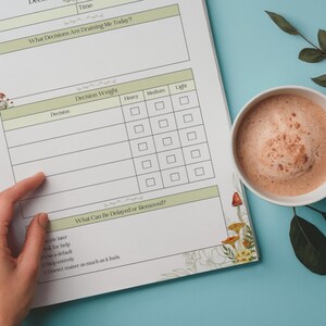 May include: A white planner with green accents and mushroom illustrations, featuring sections for decision-making and a decision weight chart. A cup of coffee sits on a blue surface. The planner includes the text: "What Decisions Are Draining Me Today?"