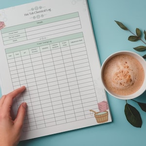 Peut inclure: Un cahier "Hot Tub Chemical Log" avec un design vert clair et blanc. Le cahier est ouvert, avec une main pointant vers la section "Water Testing Log". Une tasse de caf&eacute; et une branche de feuilles sont sur une surface bleu clair.