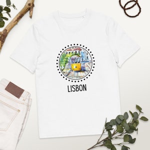 Può includere: Maglietta bianca con un'illustrazione in bianco e nero di un tram giallo a Lisbona, Portogallo. L'illustrazione è circondata da un cerchio di punti neri e la parola "LISBONA" è stampata sotto il cerchio.