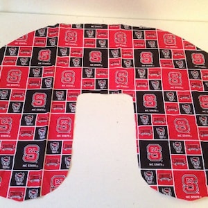 Peut inclure: Un tissu rouge et noir avec un motif répété de la lettre "N" et les mots "NC STATE". Le tissu est en forme de U et est probablement destiné à un oreiller ou un coussin.